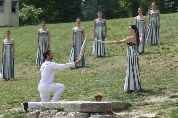 扮演最高女祭司的演員瑪麗·米娜（前右）點燃第一棒火炬手——希臘運動員、東京奧運會男子賽艇項目金牌得主斯特凡諾斯·恩圖斯科斯手中的火炬。新華社記者 趙丁喆 攝